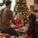 Family with a young child eating Christmas cookies together in front of a decorated Christmas tree as a holiday tradition.