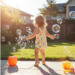 Toddler playing outside in a sunny backyard with water toys, bubbles, and sidewalk chalk on a warm summer day